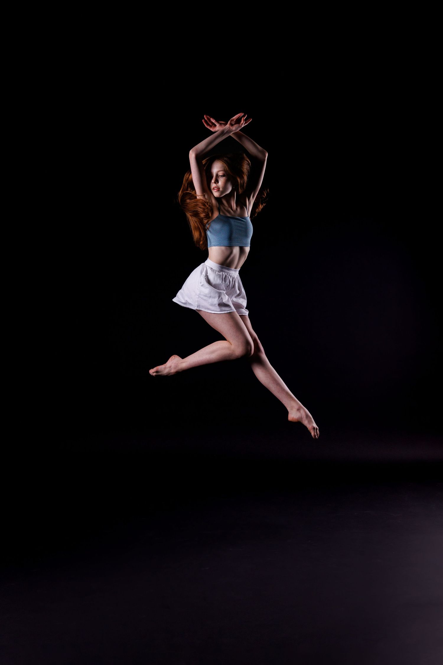 Dancer leaps gracefully with crossed arms wearing crop top and skirt on dark stage.