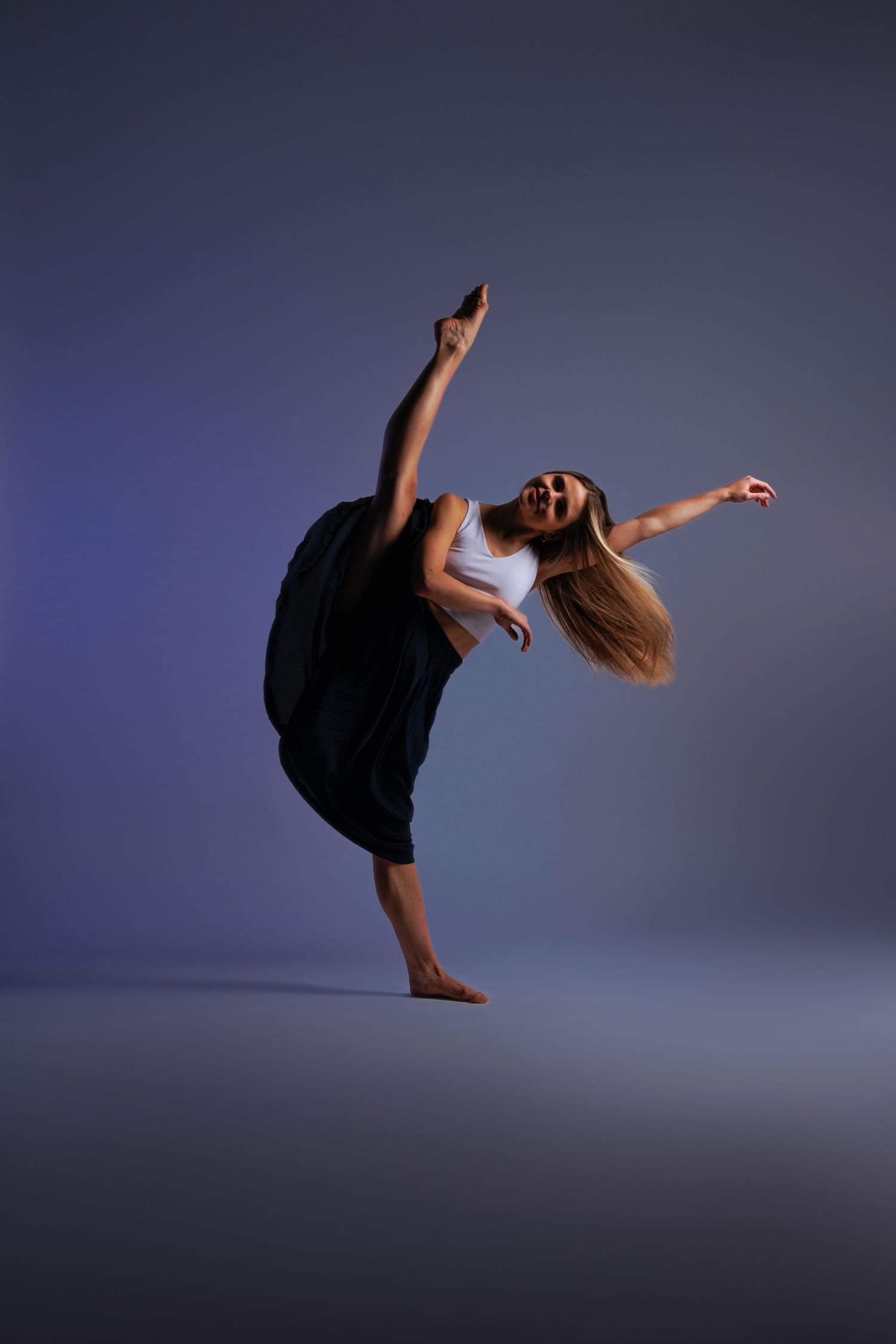 Modern dancer performing dramatic high kick in flowing black dress against purple backdrop.