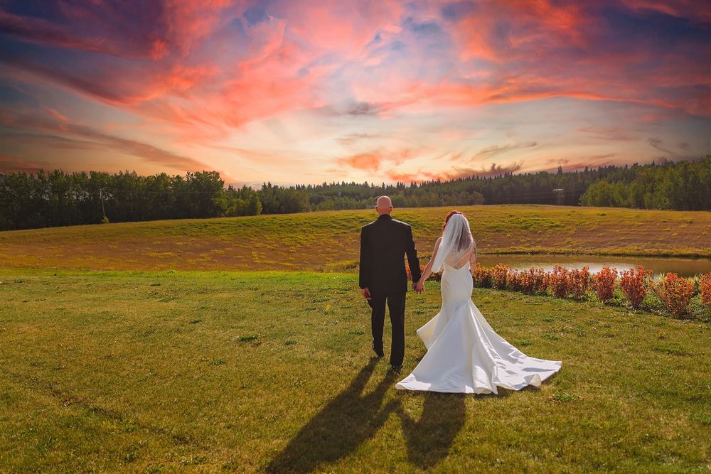 Newlyweds stand in a field during sunset with dramatic orange and pink sky.