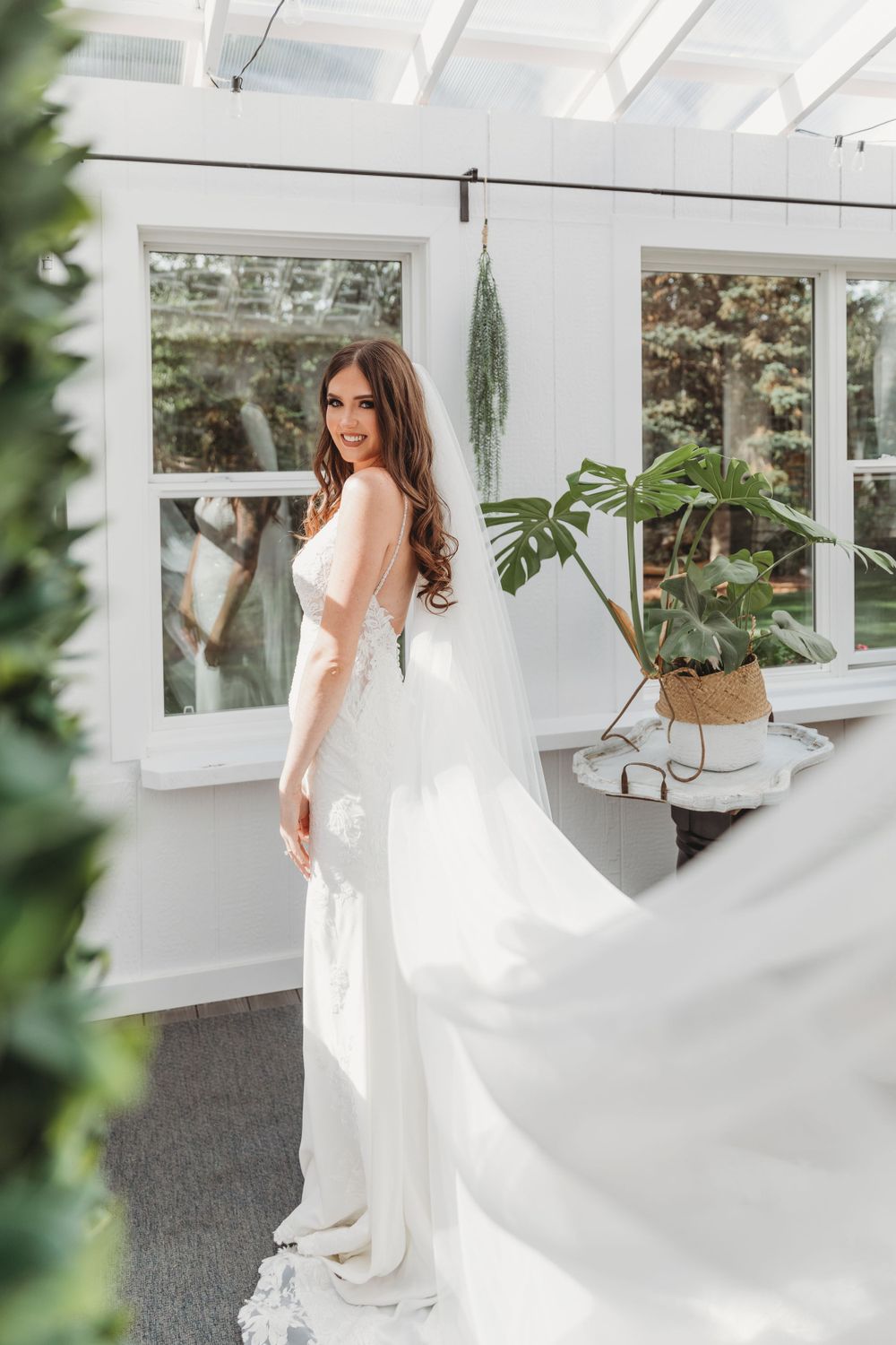 A bride in a flowing white gown poses in a sunlit conservatory with indoor plants.