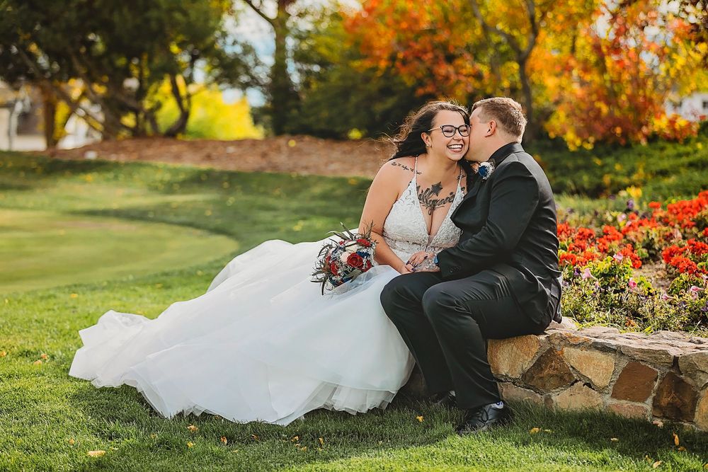 Romantic wedding couple sits on stone wall amid autumn flowers and green lawn.