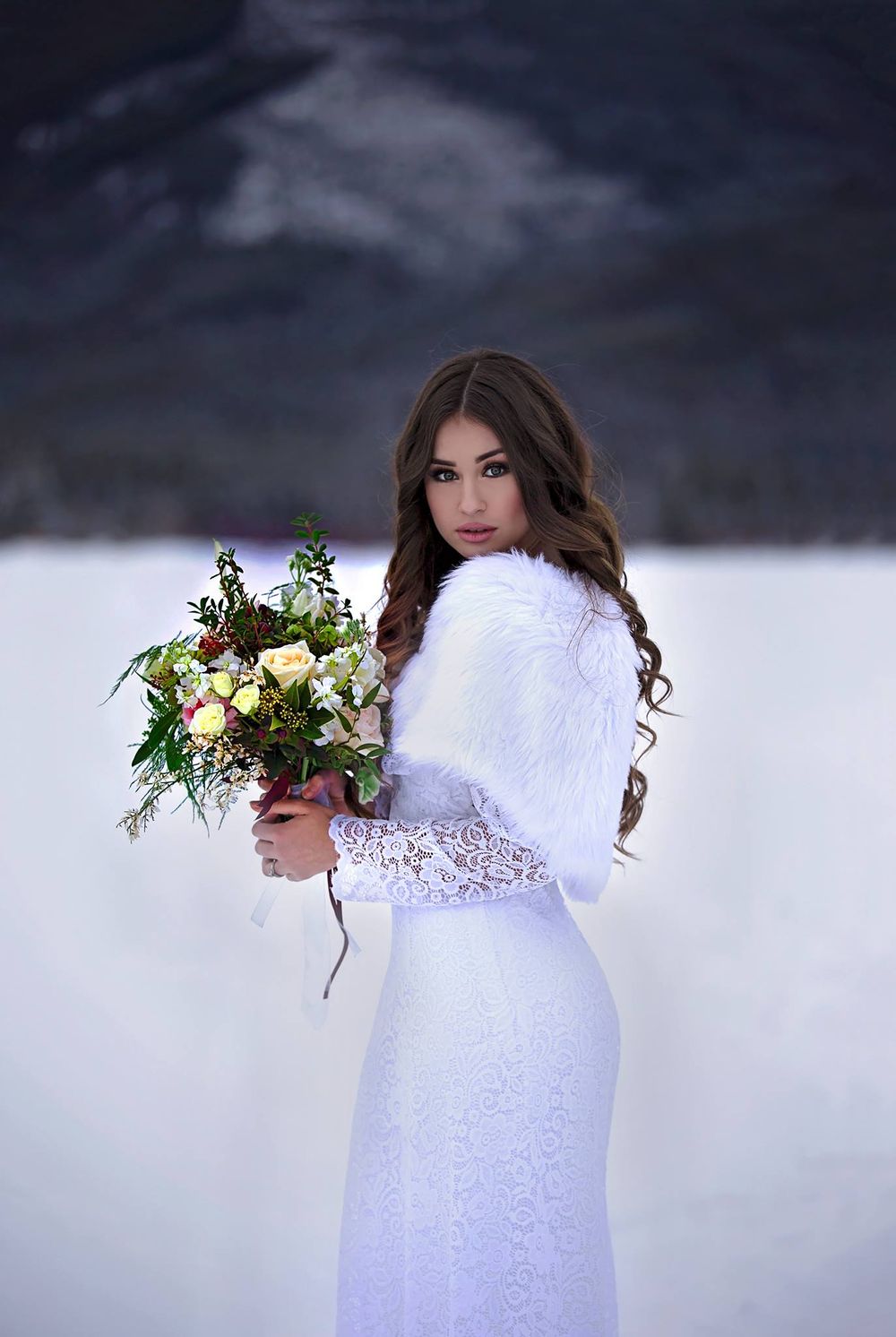 Winter bride in white lace dress holds bouquet while posing in snowy landscape.