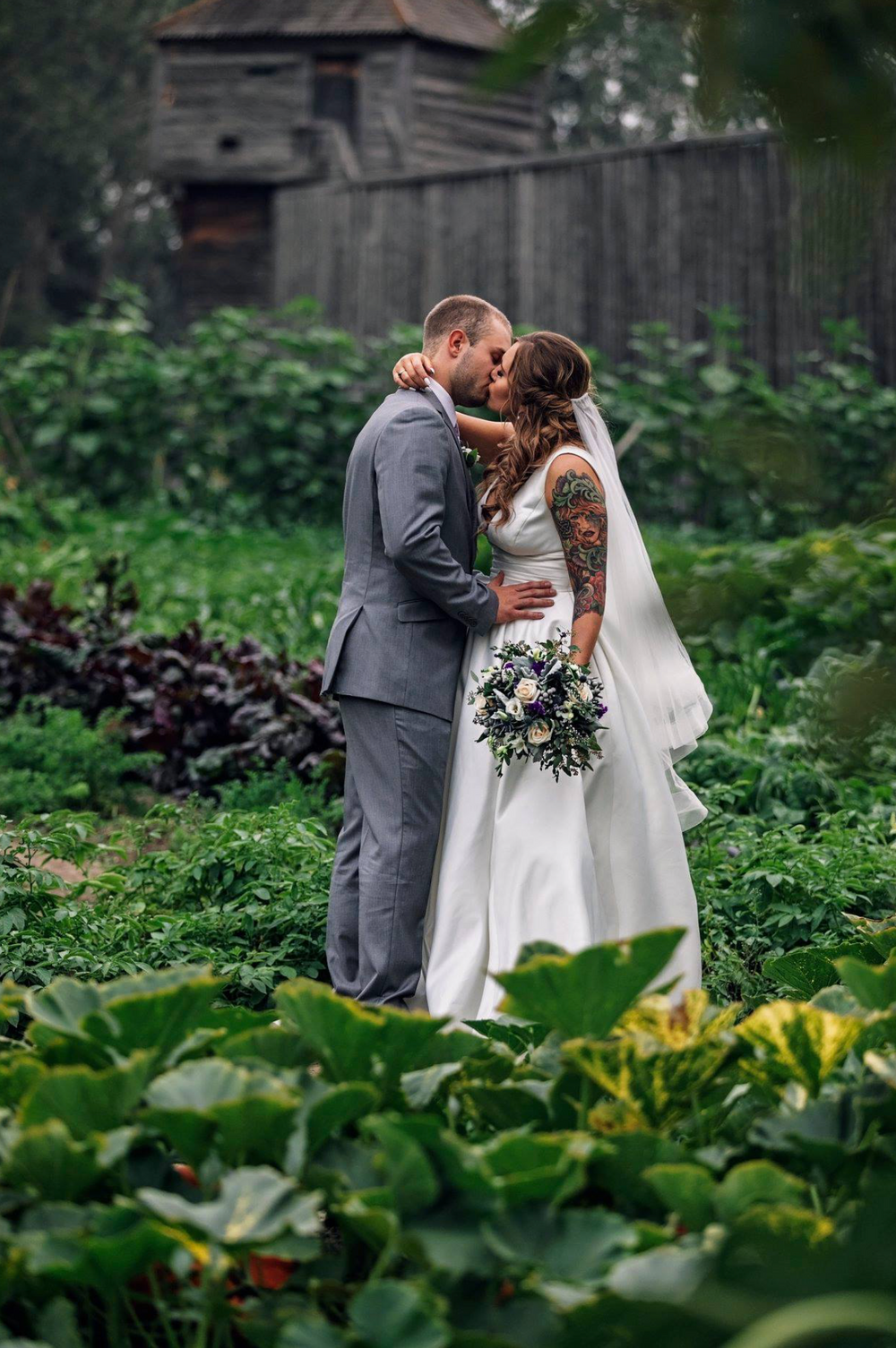 Newlyweds share a romantic kiss in a lush garden setting next to a rustic barn.