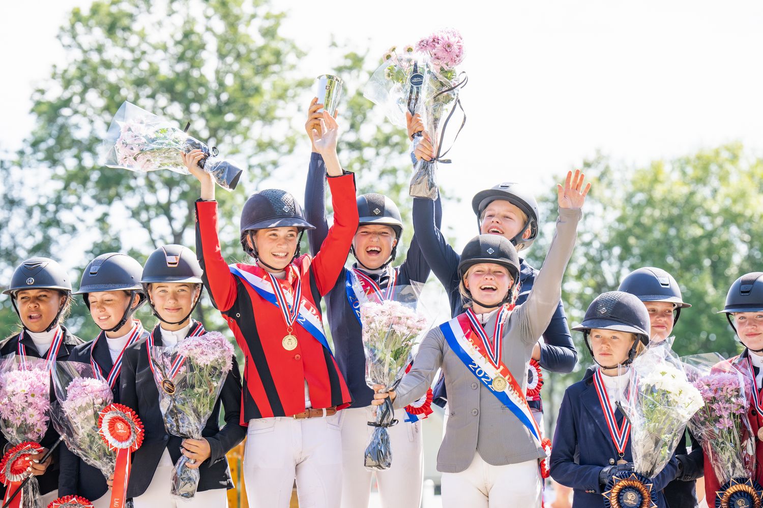 Group of equestrian competitors celebrating with trophies and flowers at awards ceremony.
