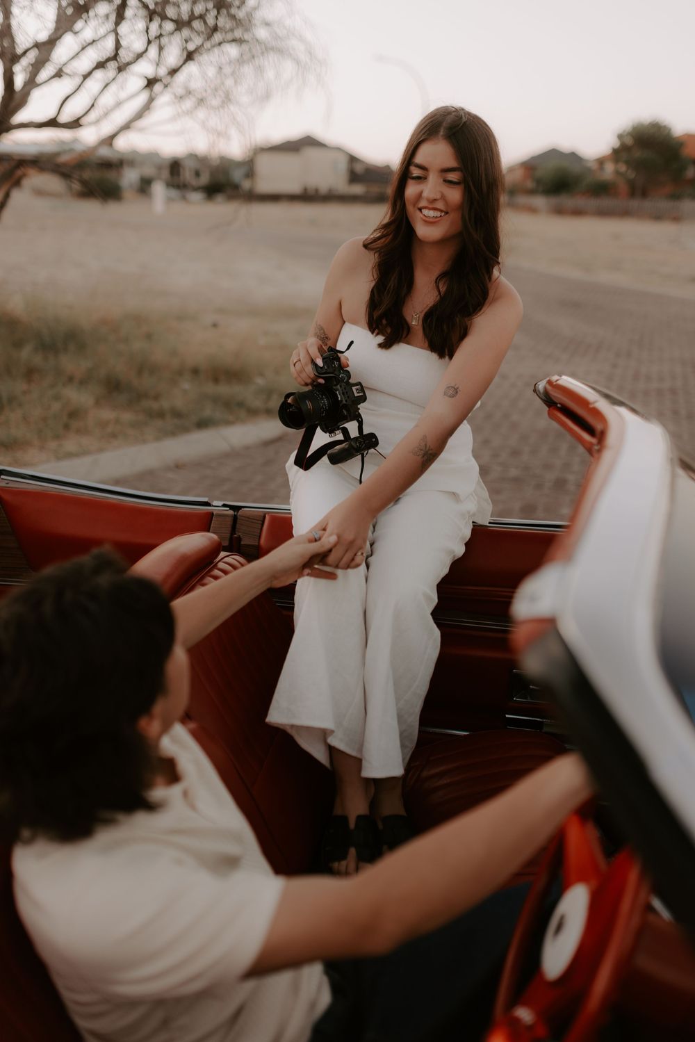 A woman in a white dress smiles while being helped into a vintage red convertible car at sunset.