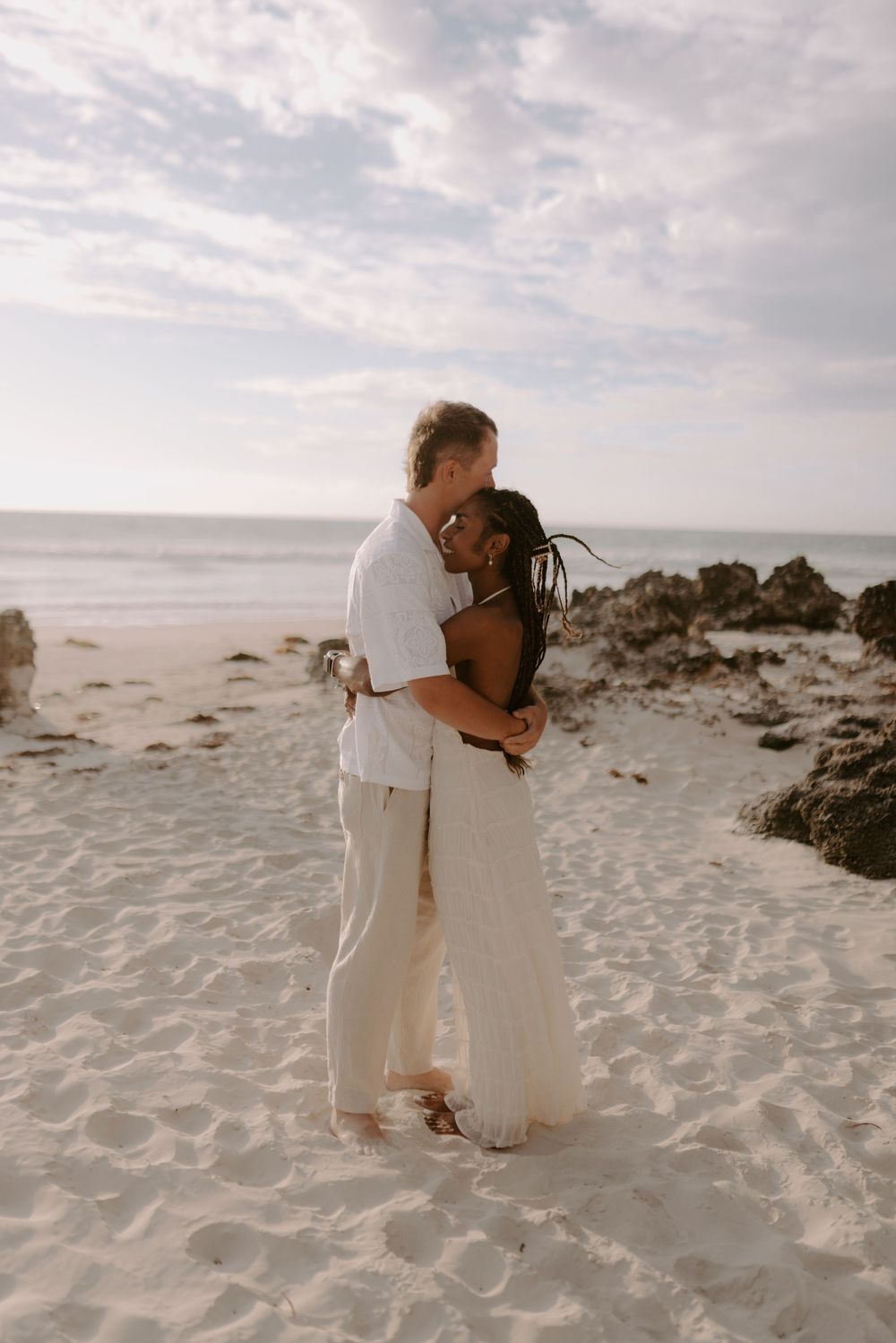 A romantic couple embraces on a serene white sandy beach with cloudy skies and rocky outcrops in the background.