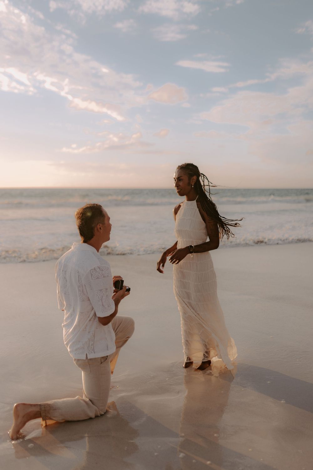 Two people in white clothing share a romantic proposal moment on a sunny beach at sunset.
