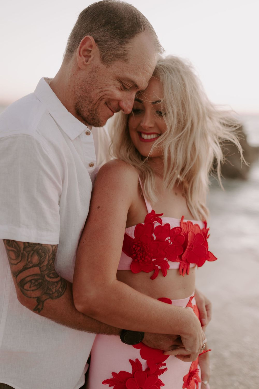 Couple shares playful moments on beach, woman wearing vibrant red floral dress.
