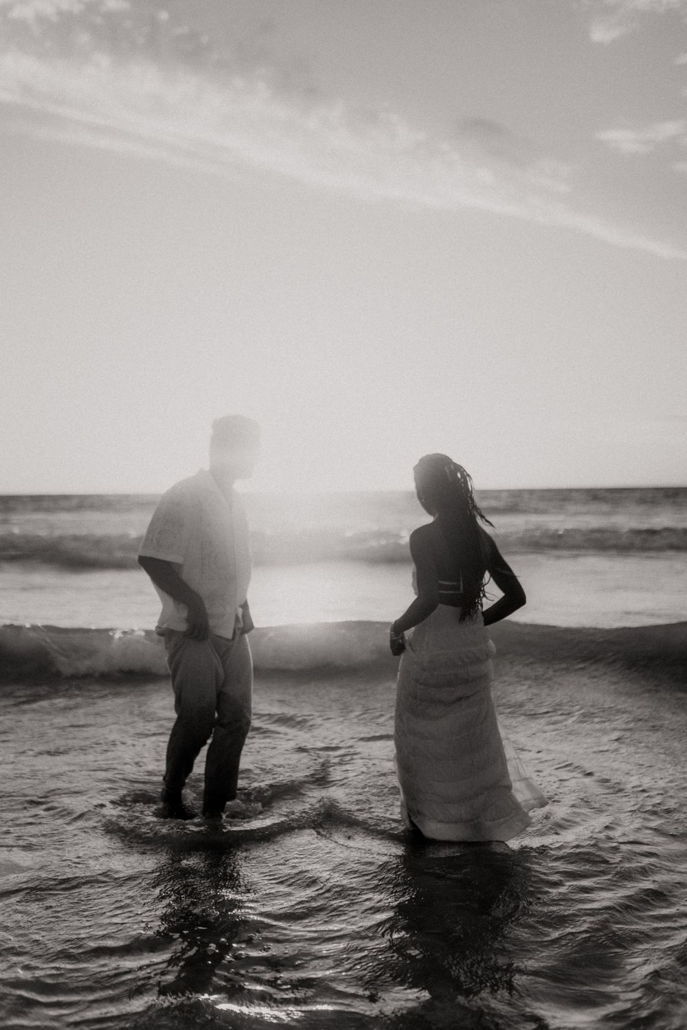 A silhouetted couple stands in shallow ocean waves at sunset creating a romantic black and white beach scene.