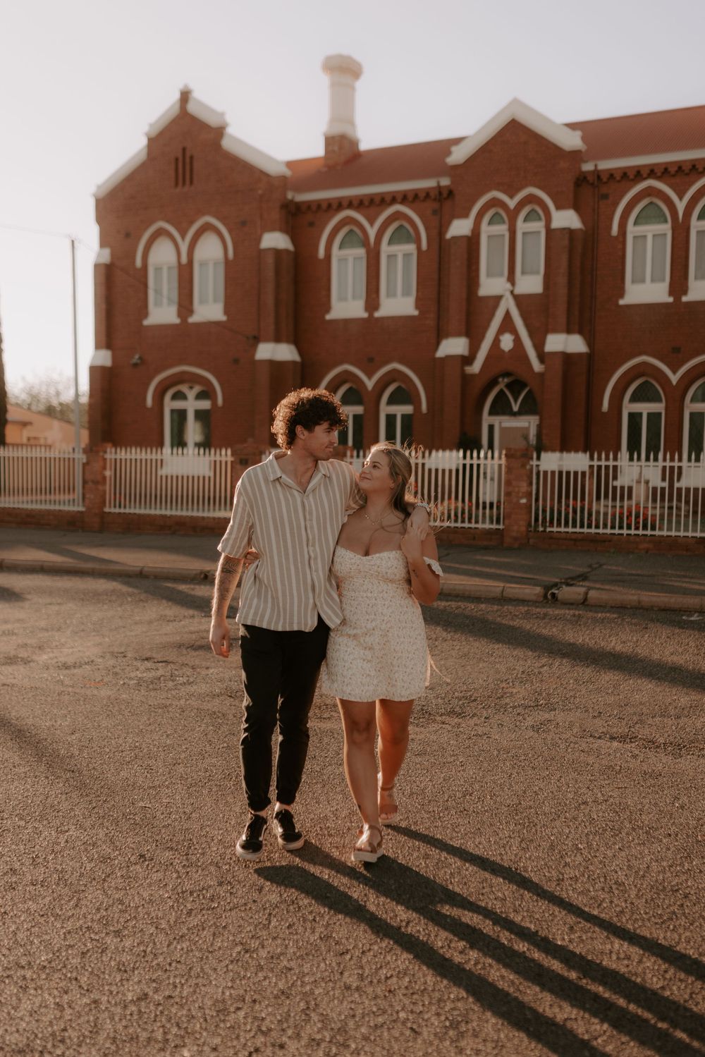 A couple walks hand in hand casting long shadows against a brick building backdrop during golden hour.