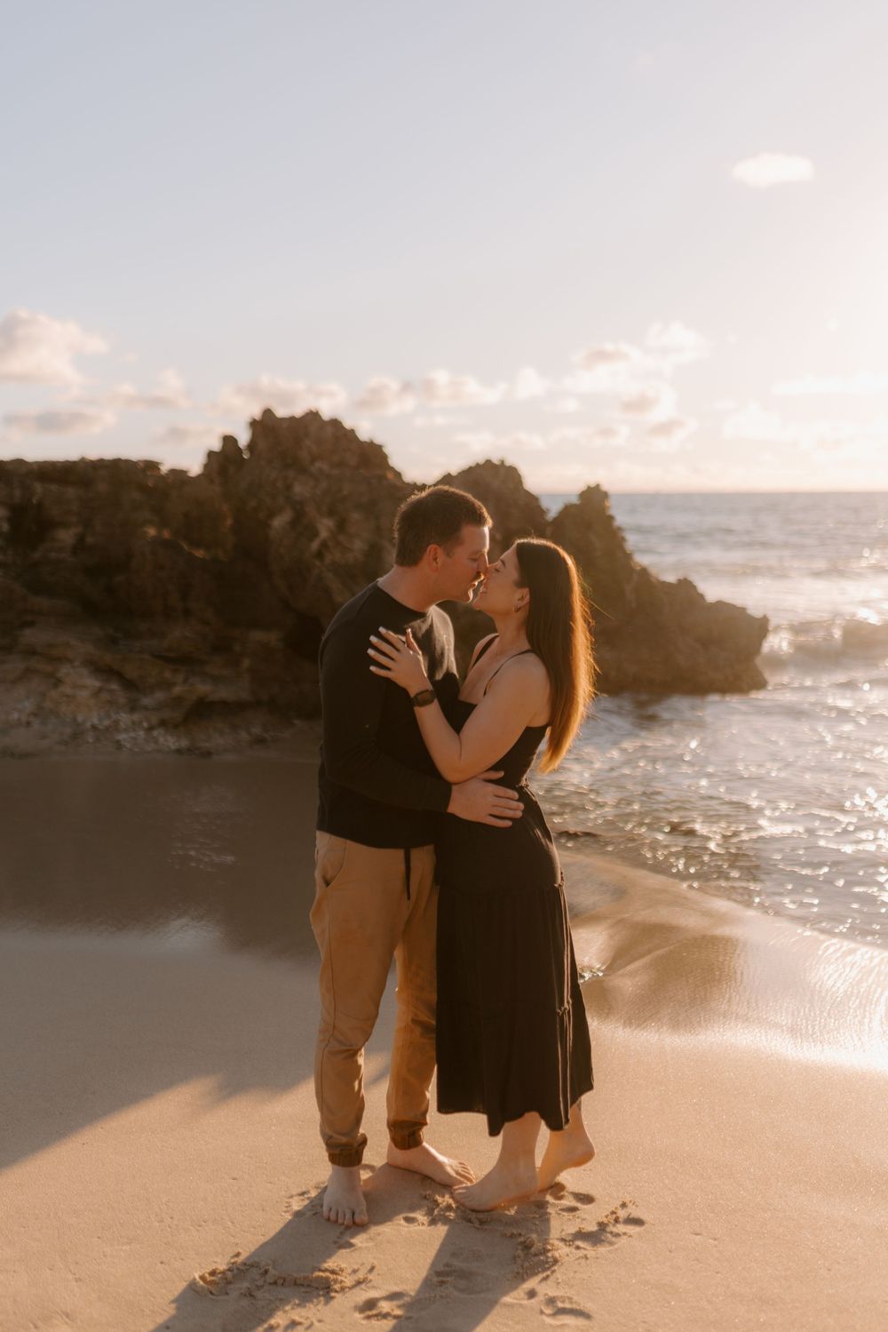 Romantic couple embracing on a sandy beach at sunset with rocky coastline and ocean waves in the background.