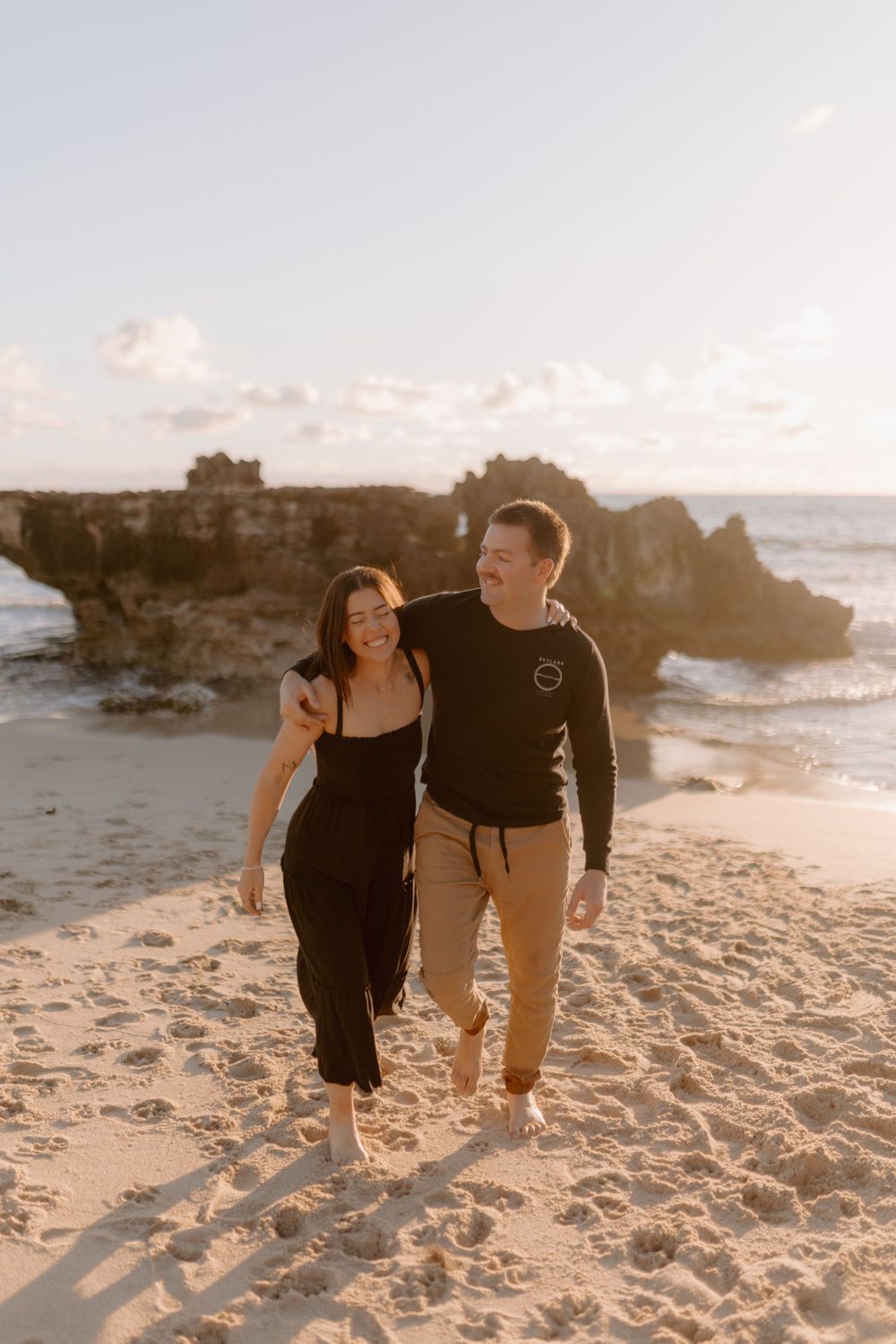 Couple walking together on a beach at golden hour, with dramatic rocky cliffs and sparkling ocean views.