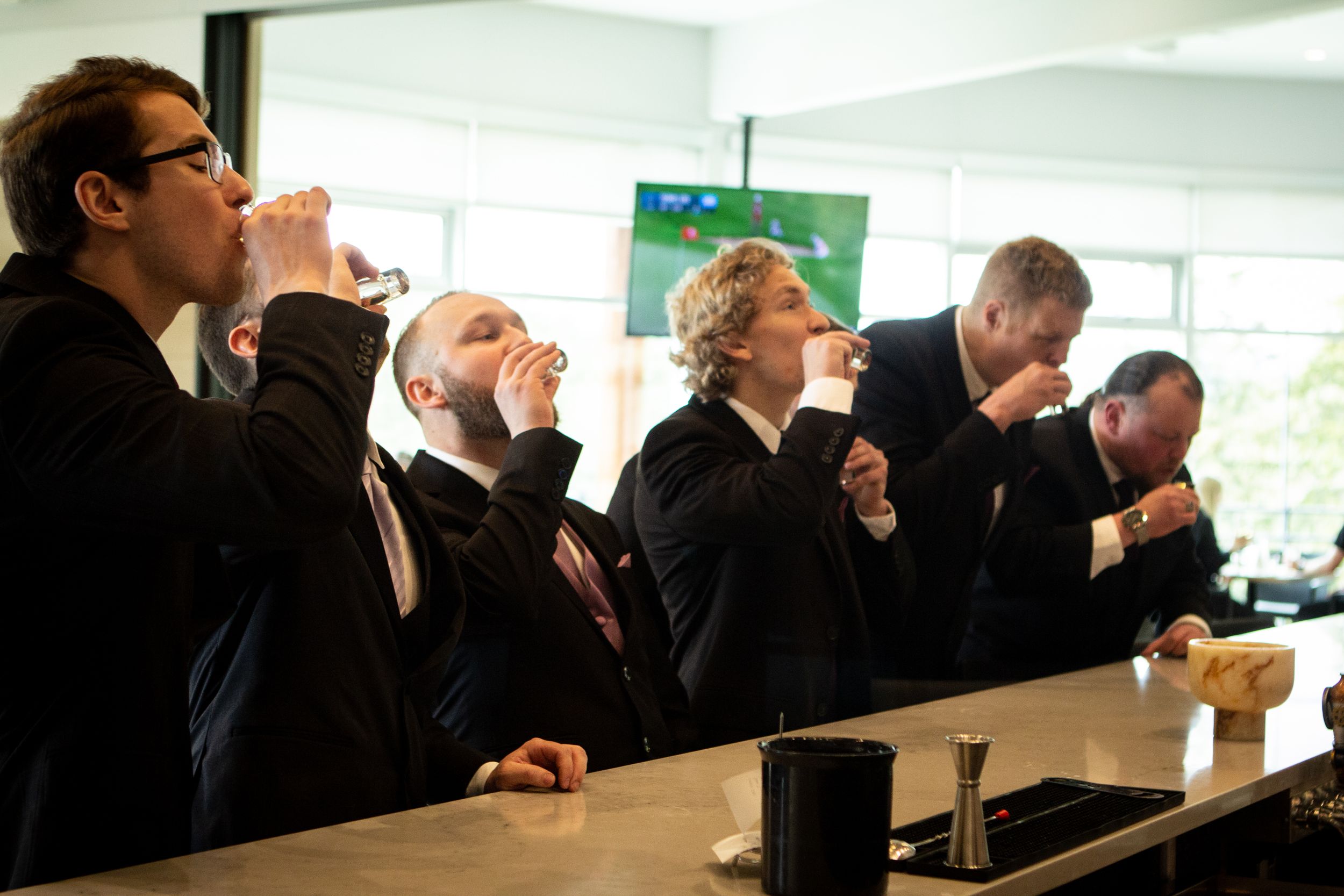 A group of groomsmen taking shots at a bar counter during a wedding celebration.