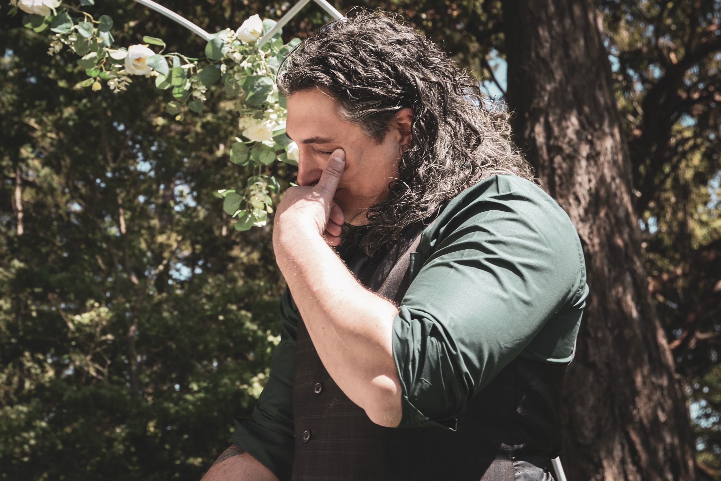 Person with curly hair and green jacket stands in wooded area while holding a camera to their face.