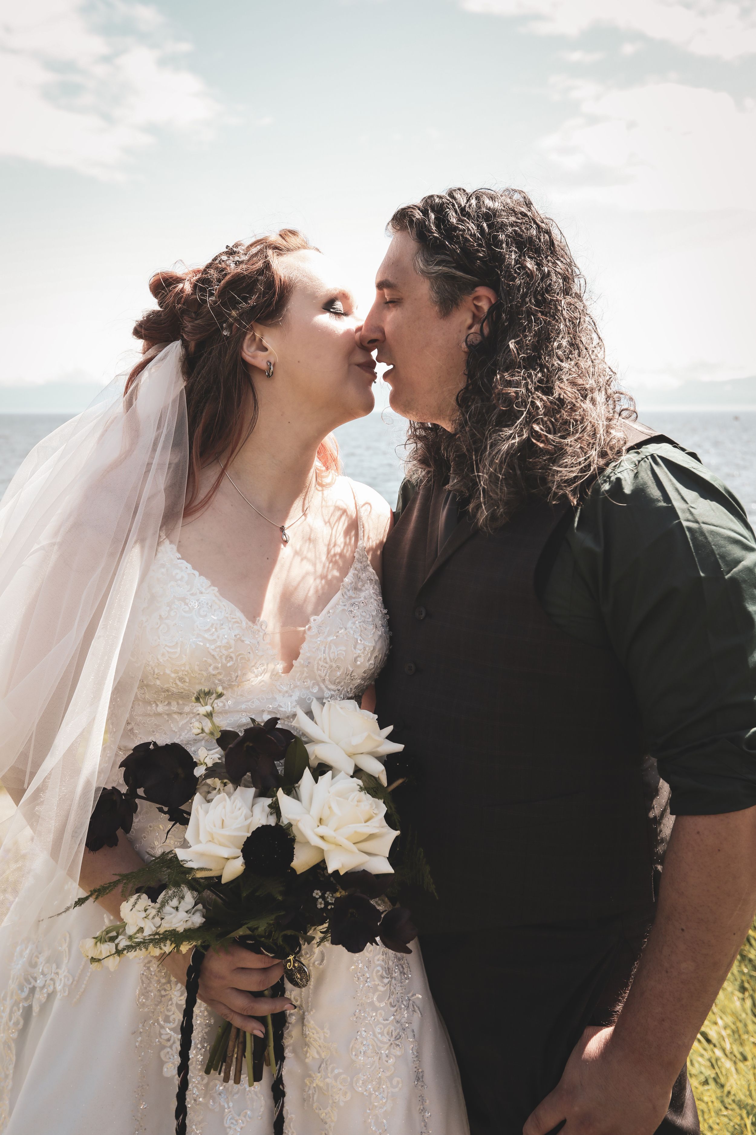 A romantic wedding couple shares a tender moment outdoors with a bouquet of black and white roses against a cloudy sky.