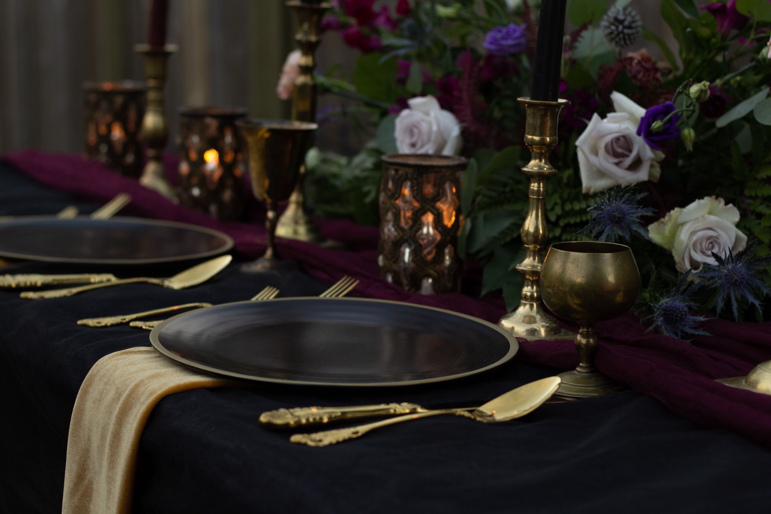 An elegant dark table setting with gold cutlery, lanterns, and purple roses creating a romantic ambiance.