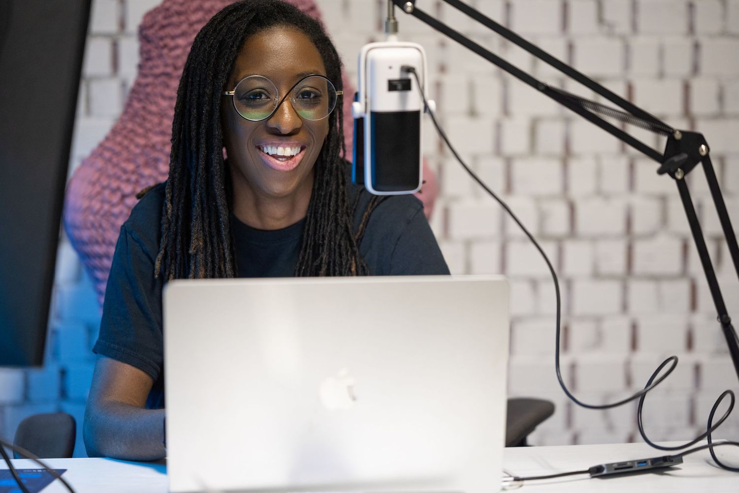 Content creator with long braids smiling while recording podcast at desk with laptop and microphone.