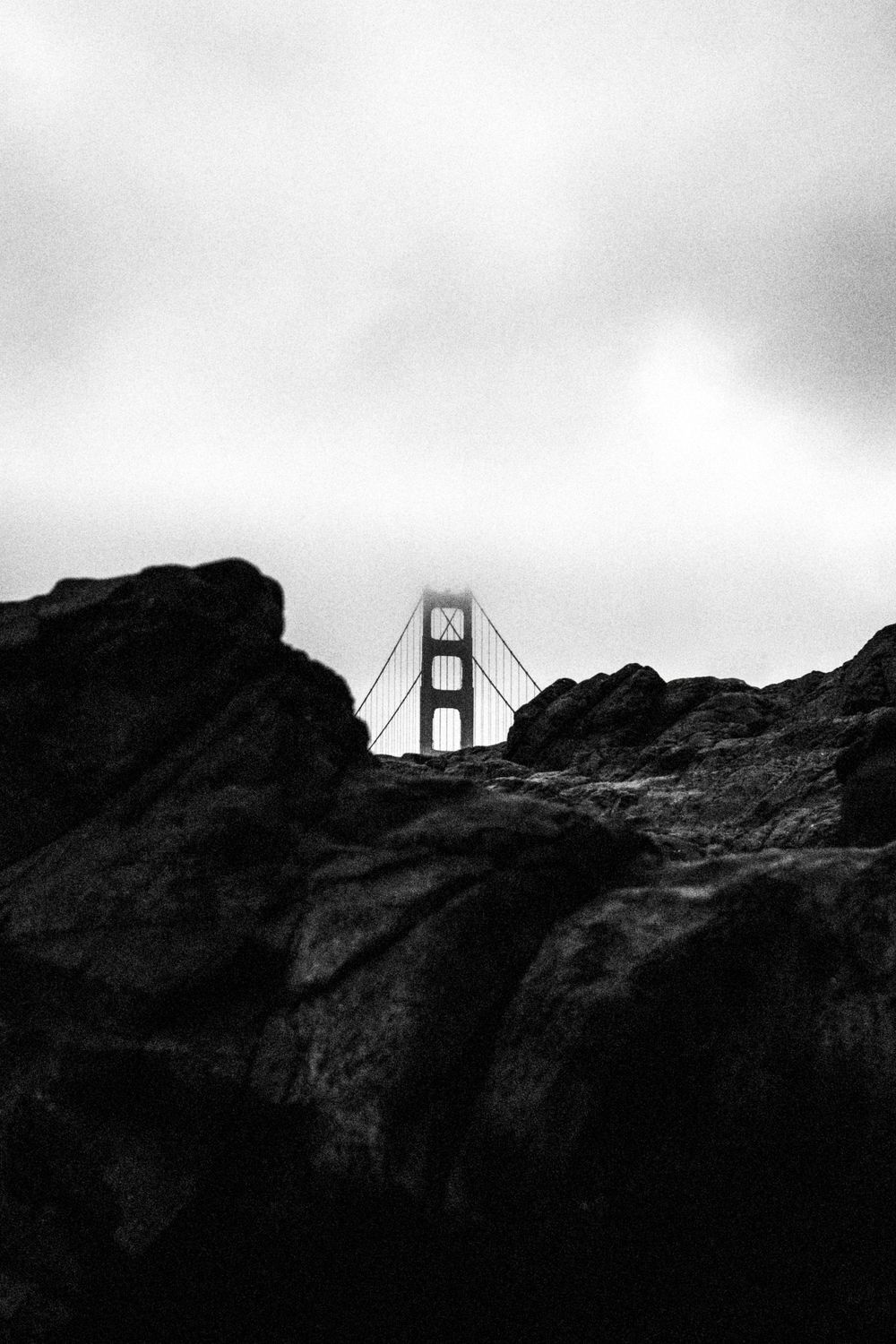 Moody black and white photograph of Golden Gate Bridge peeking through rocky foreground.