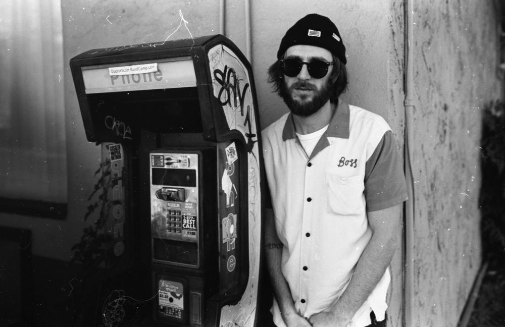 Person in casual attire standing next to retro payphone in black and white photography.