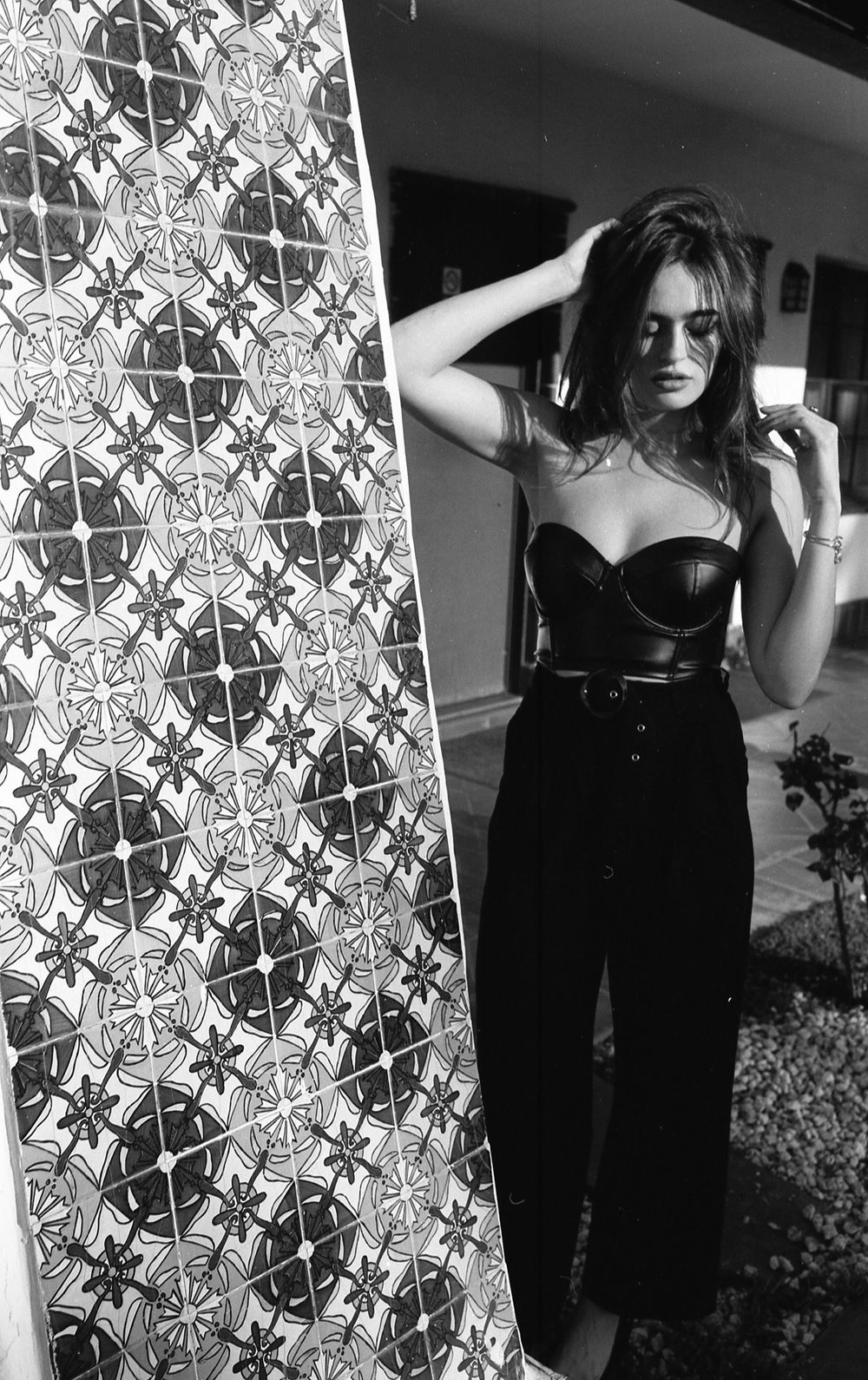 Black and white portrait against patterned wallpaper featuring stylish dark outfit.