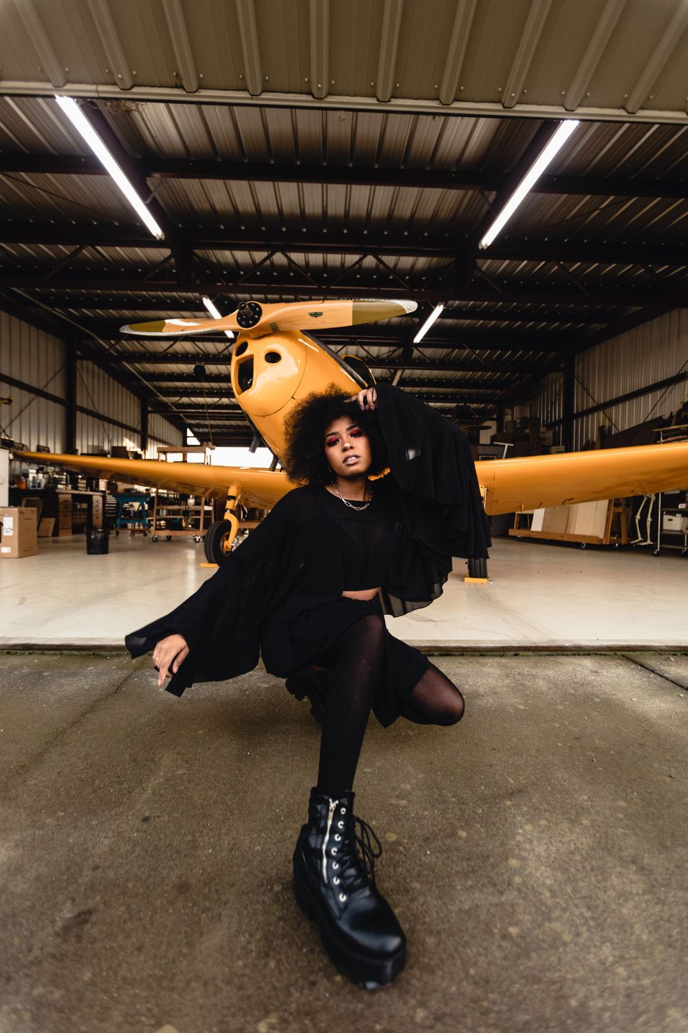 Person posing playfully in black outfit in front of vintage yellow airplane in hangar.