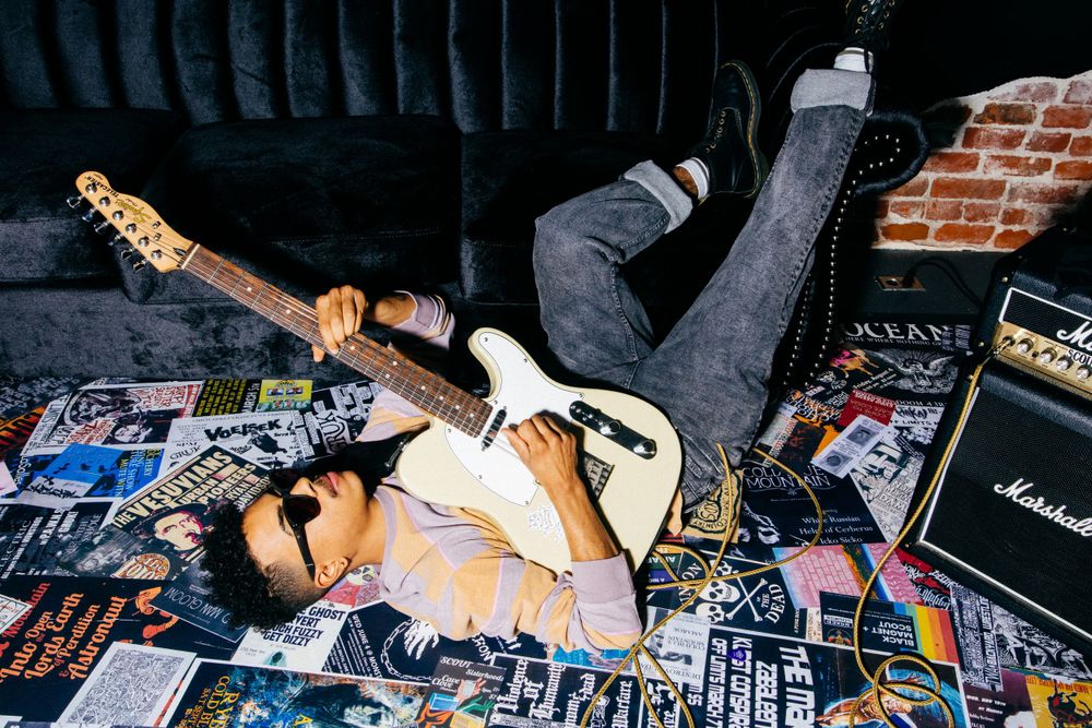 Musician lying on floor covered in magazine clippings with Fender guitar and Marshall amp.