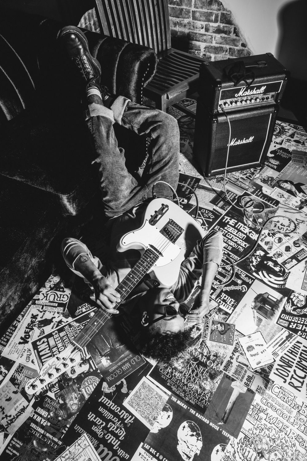 Black and white photo of a musician lying on floor covered in posters with electric guitar and Marshall amps.