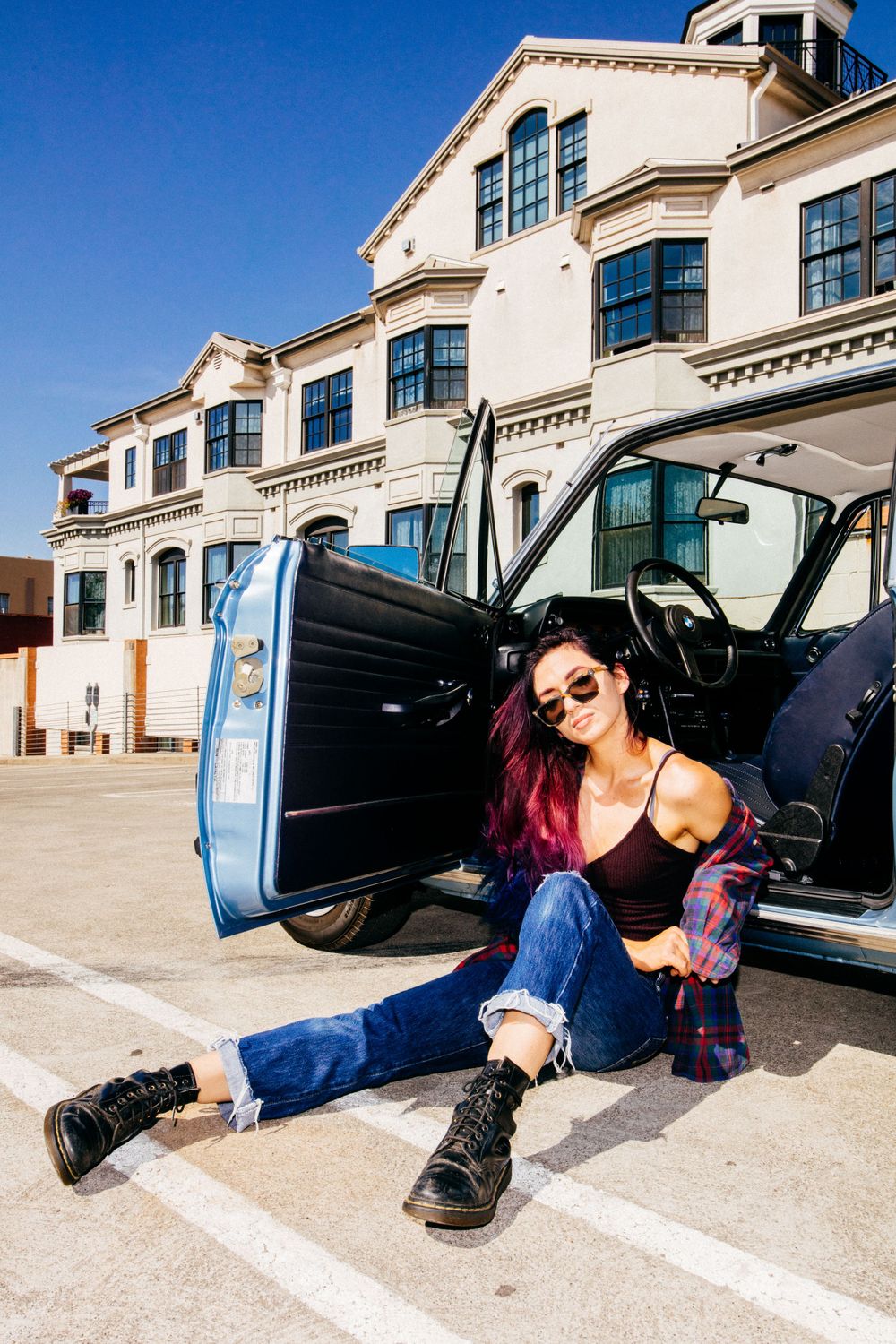 Stylish car photography featuring distressed blue muscle car and casual streetwear fashion against urban building backdrop.
