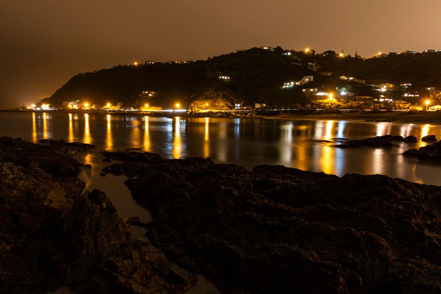 Golden lights ripple across Wellington Harbour, casting a warm glow over the quiet shoreline.