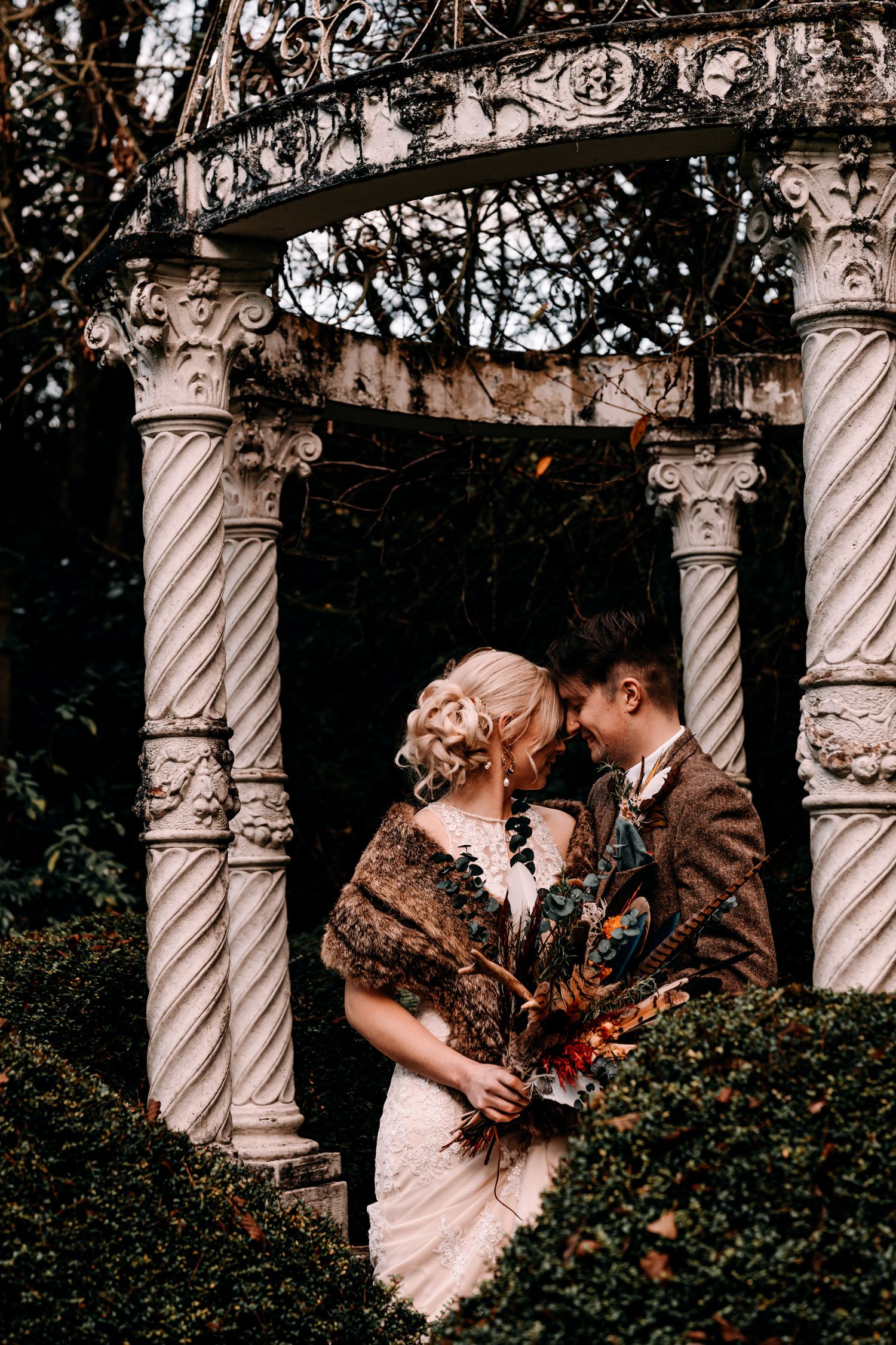 A couple in vintage wedding attire share an intimate moment under ornate stone columns in a garden setting.