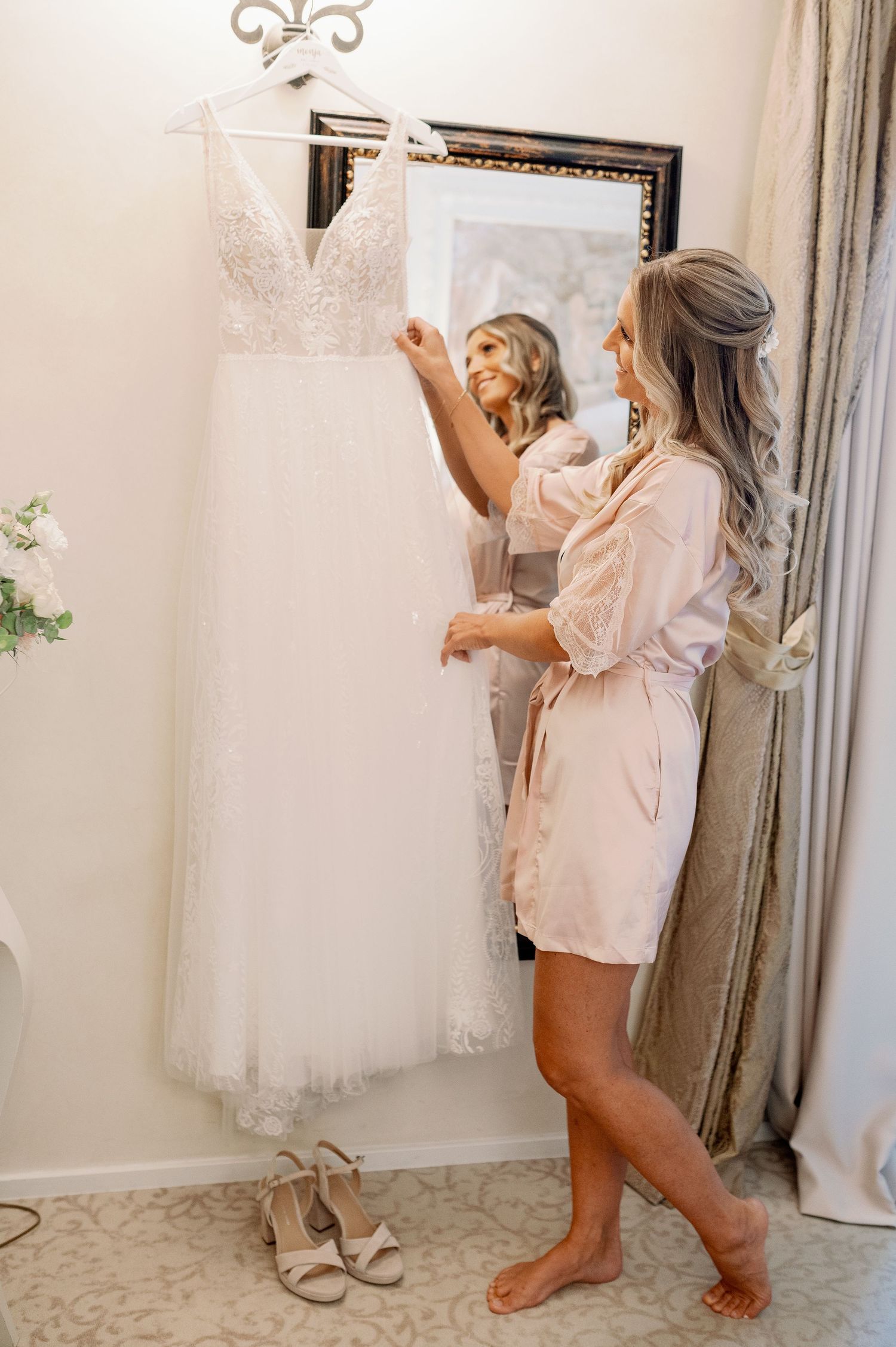 A white lace wedding dress hangs on display while someone in a blush robe admires it in a bridal suite.