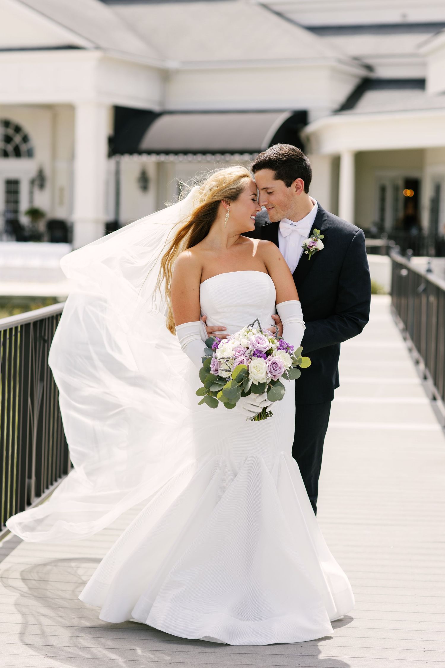Elegant bride and groom share intimate moment on walkway with flowing veil and purple bouquet.