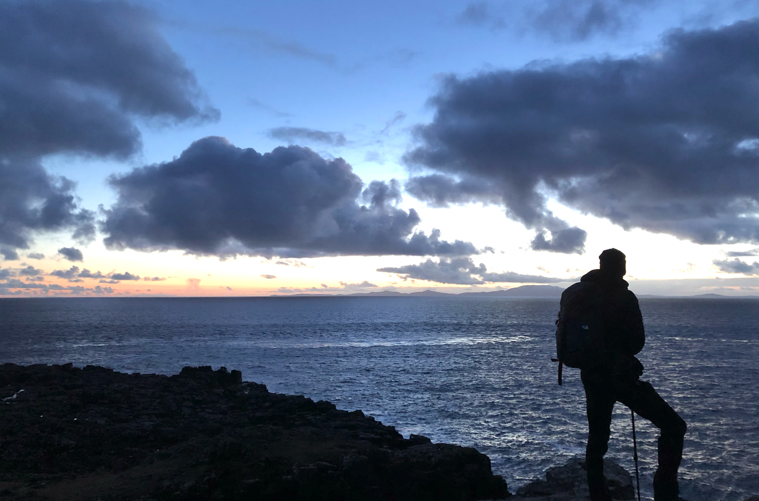 Silhouette of person standing on cliff edge overlooking dramatic ocean sunset.