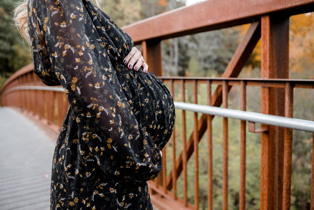 Woman in patterned black dress poses on rustic wooden bridge with railing.
