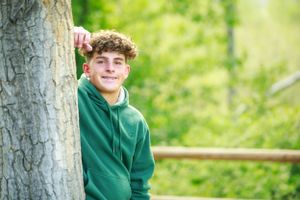Youth in green hoodie leaning against tree trunk in sunny outdoor setting.