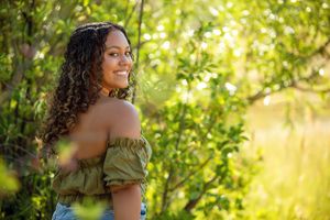 Sun-drenched portrait through green foliage wearing an olive-colored top.