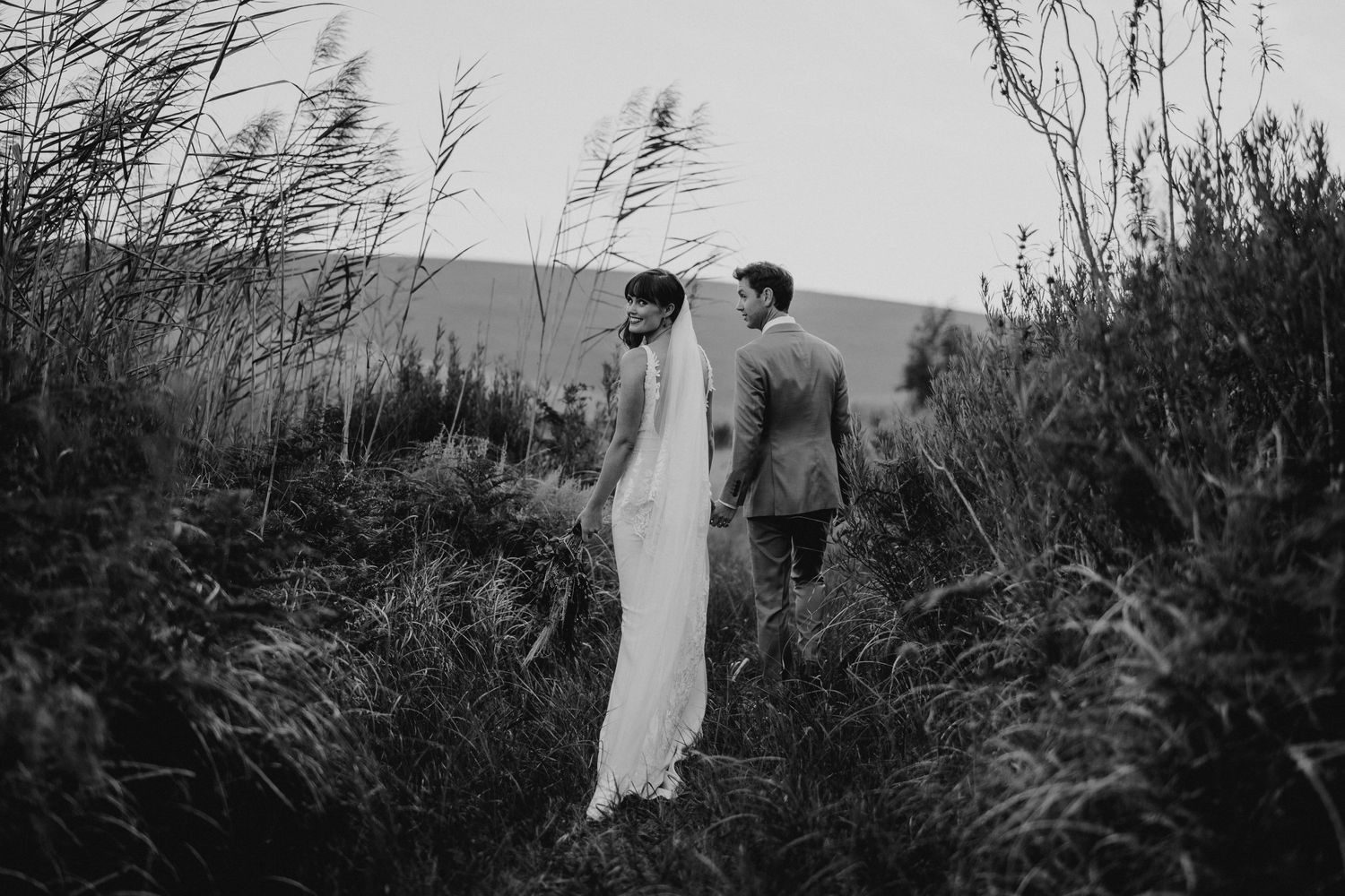 Black and white portrait of couple in wedding attire among tall grasses.