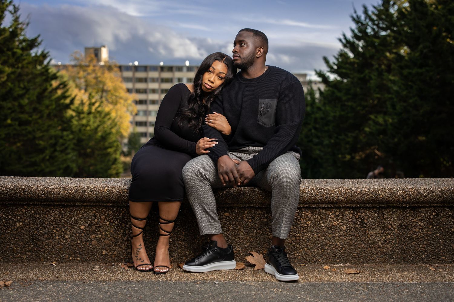Couple sharing intimate moment while sitting on stone wall during sunset with cityscape in background.