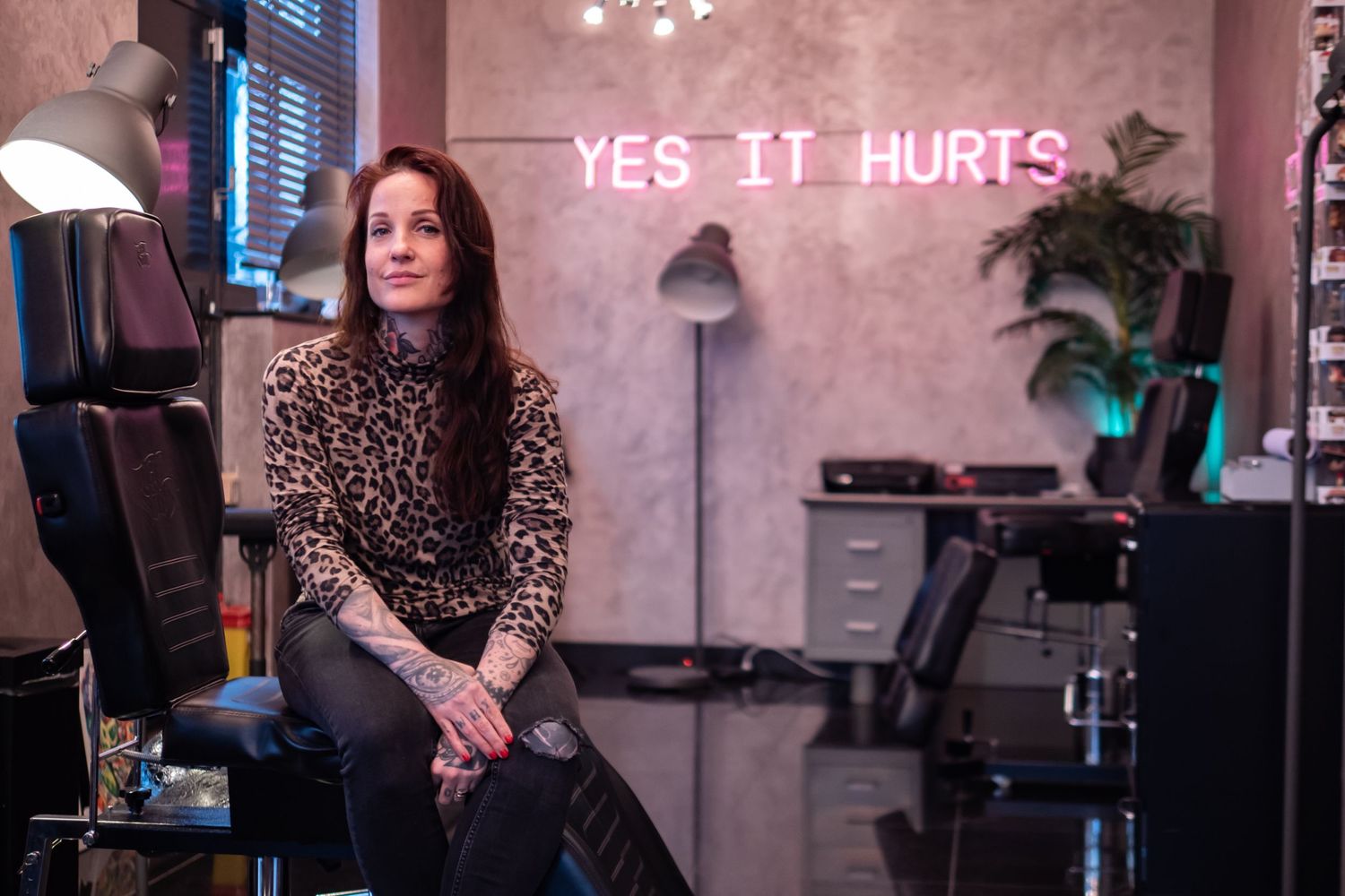 Tattoo shop owner sitting in her shop in front of a sign that says "yes it hurts".