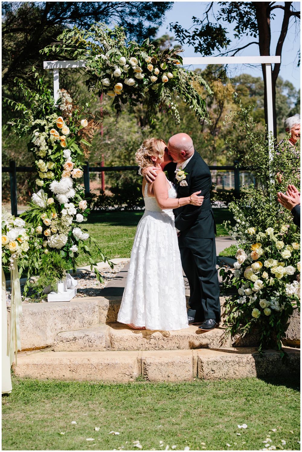 Wedding ceremony under a romantic arch adorned with peach and cream roses in garden setting.