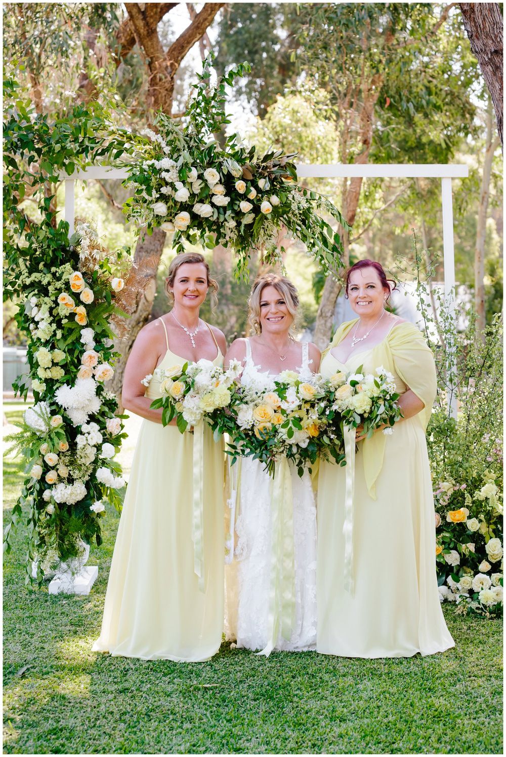 Three bridesmaids in pale yellow dresses pose under floral arch at outdoor garden wedding.