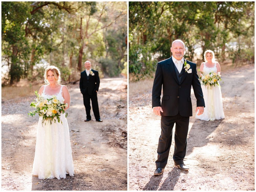 Bride in lace gown poses on wooded path while groom walks towards her in black suit.