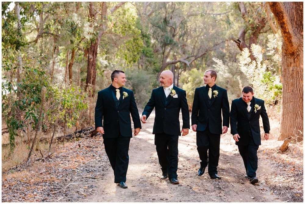 Four groomsmen in black tuxedos walking together down a forest path.