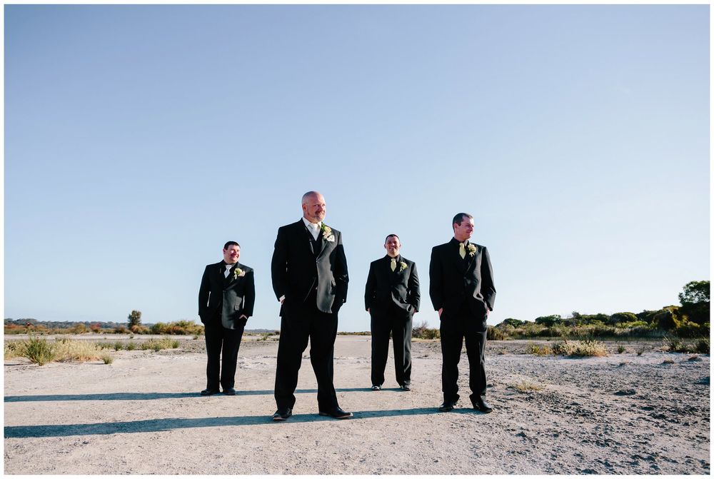 Groomsmen in black suits stand against bright blue sky in desert landscape.