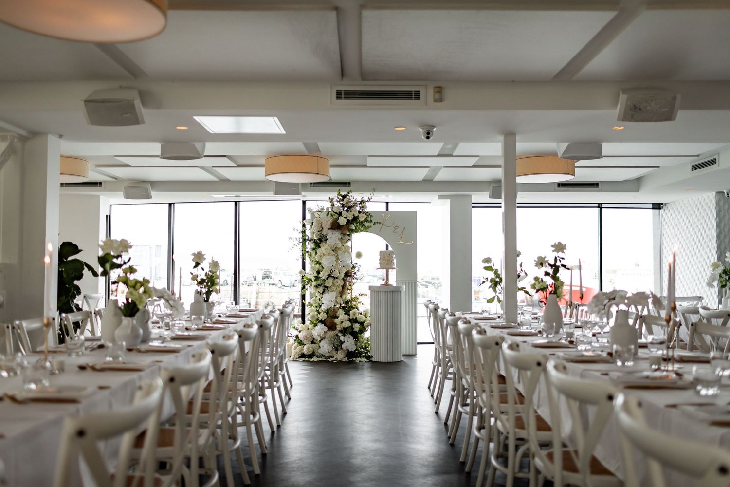 Light-filled modern wedding reception venue with white tables and floral arrangements.