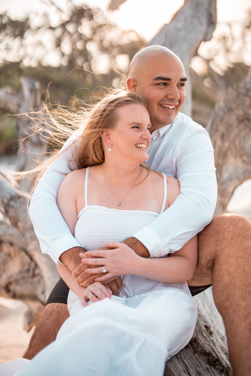 Intimate beach portrait of couple sharing laughter while sitting on driftwood.