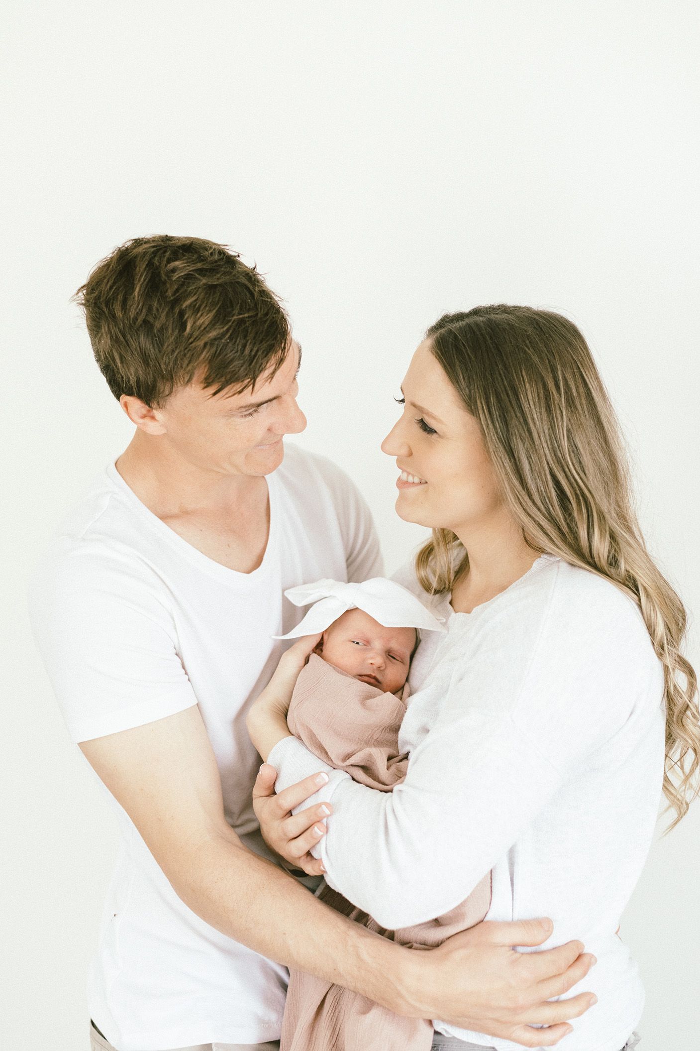 Intimate family portrait with parents holding newborn against white backdrop.