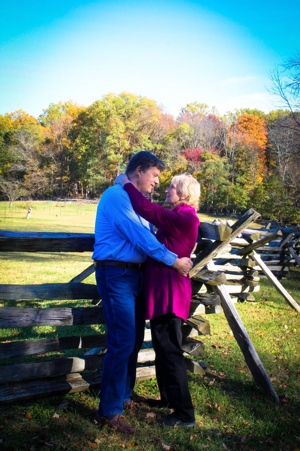 A couple embraces near a wooden fence during autumn with colorful foliage.