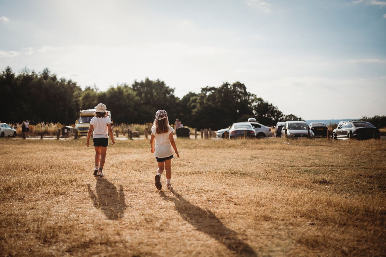 Two people walking across a dry summer field with parked vehicles in the background.