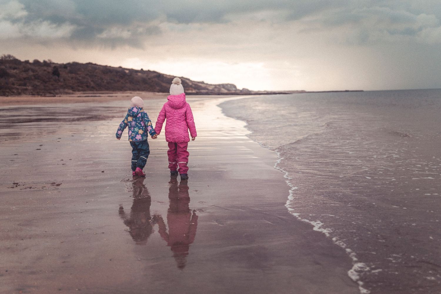 Two bundled-up figures walking along a misty beach at sunset with their reflections in the wet sand.